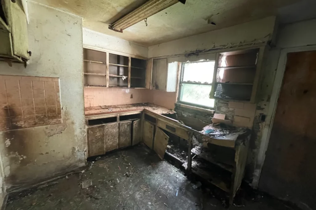 Damaged kitchen interior showing soot-covered walls, broken wooden cabinets, peeling paint, and debris on the floor in Jacksonville, FL.