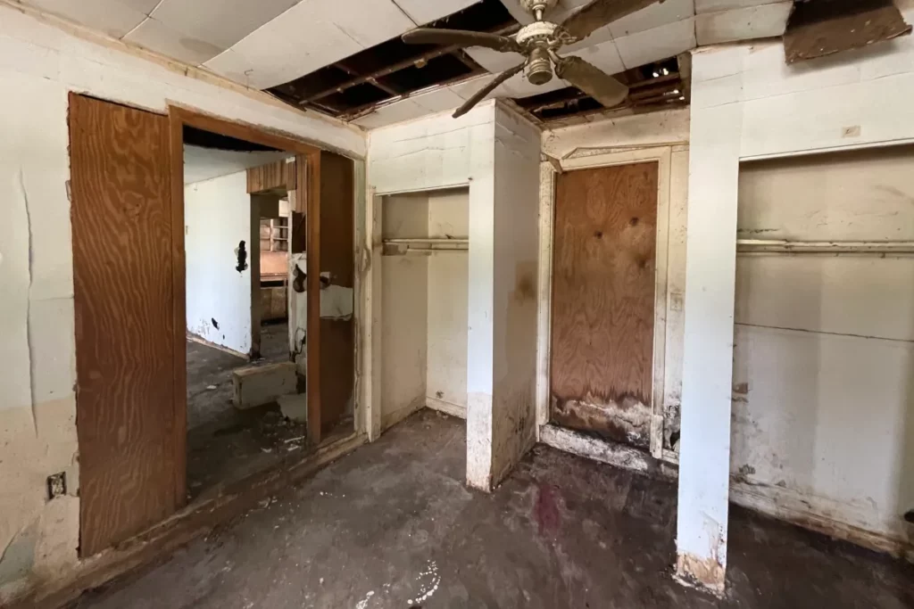 Unfinished master bedroom space showing water-damaged drop ceiling, exposed wooden framing, plywood doorway, and bare concrete floor in Jacksonville, FL.