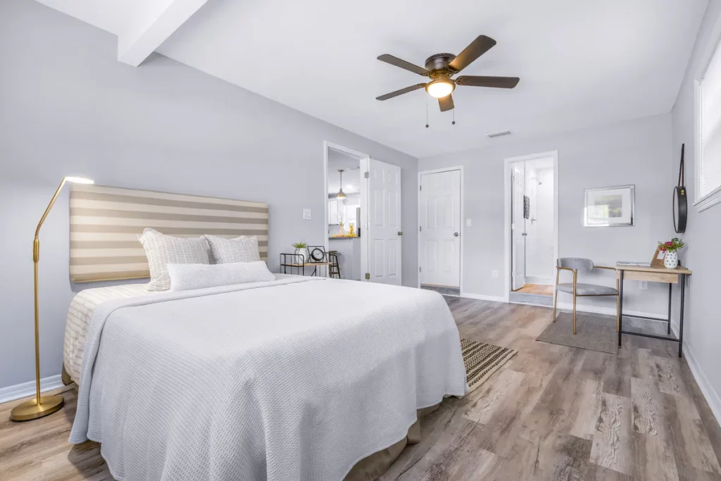 Residential bedroom interior displaying a bed with striped headboard, grey painted walls, wood-grain flooring, ceiling fan, standing floor lamp, and writing desk in Jacksonville, FL.