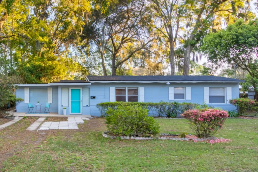Exterior view of a single-level light blue residential house with a bright turquoise door, white shutters, front lawn landscaping, and oak trees in Jacksonville, FL.
