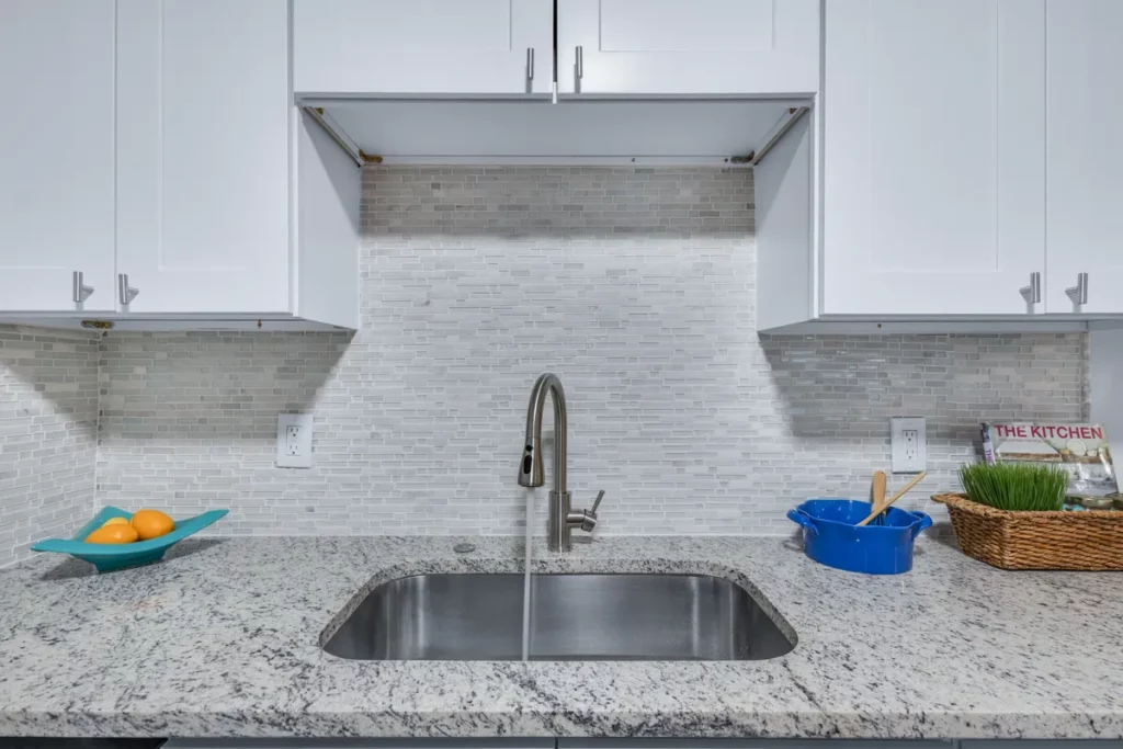 A single-basin stainless steel undermount sink with running water, light granite countertops, white shaker cabinets, and a narrow stone mosaic backsplash in Jacksonville, FL.