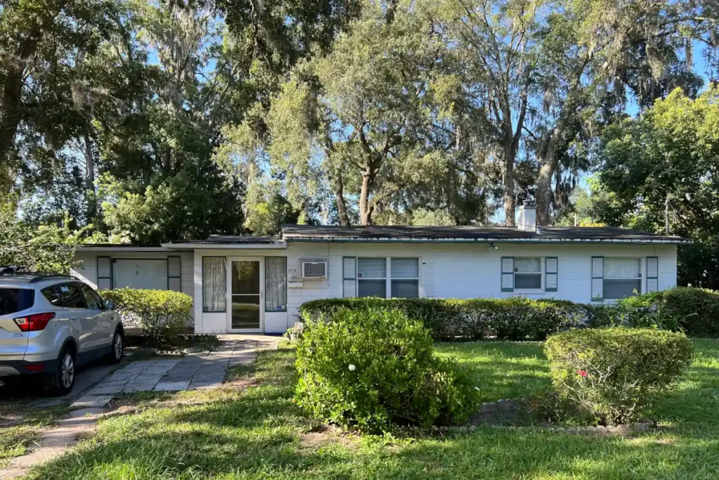 Exterior view of a white single-level house with dark green shutters, a front storm door, a lawn with manicured shrubs, and mature oak trees in Jacksonville, FL.