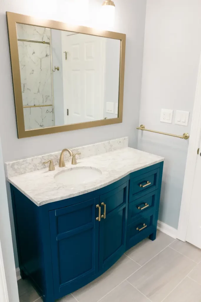 Navy blue single-sink bathroom vanity with brass faucet, marble-look countertop, gold mirror frame, and light grey tile floor in Jacksonville, FL.