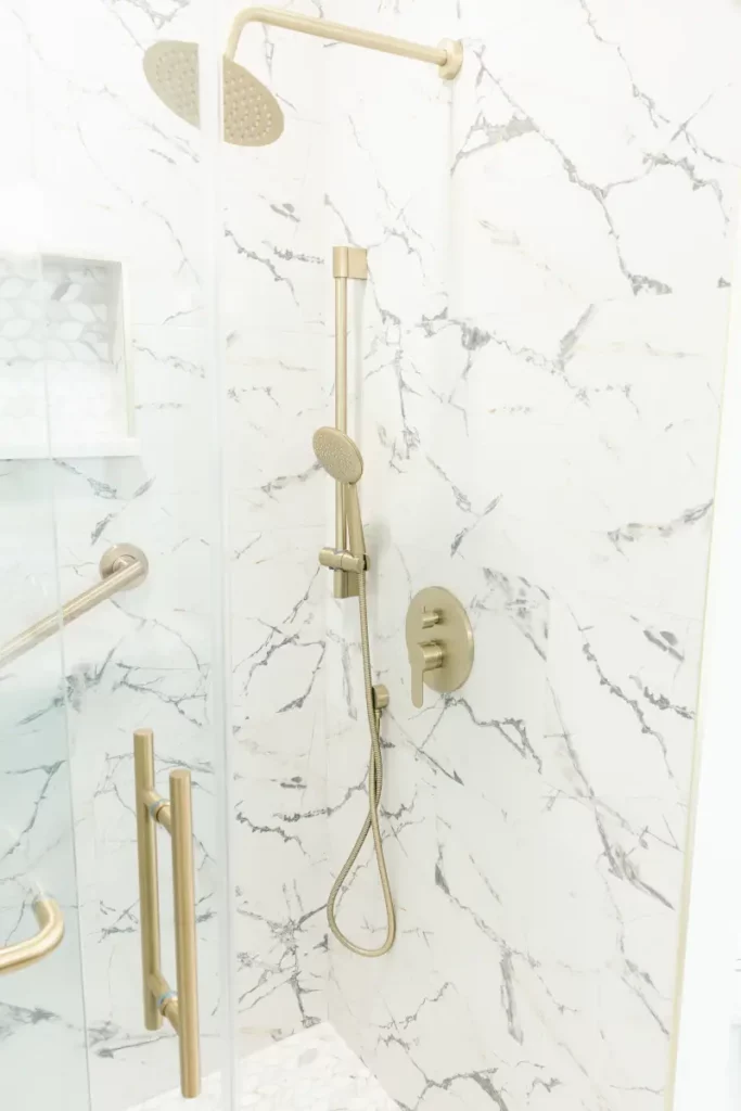 Interior view of a glass-enclosed shower featuring white and grey calacatta marble wall tiles, brushed brass rain head, handheld wand, and grab bar in Jacksonville, FL.