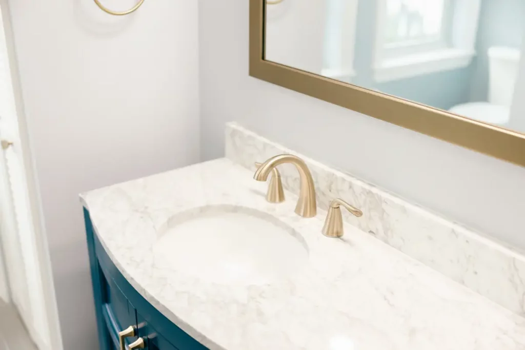 Close-up view of an oval undermount bathroom sink, white marble counter, gold three-piece faucet fixture, and metallic framed mirror in Jacksonville, FL.