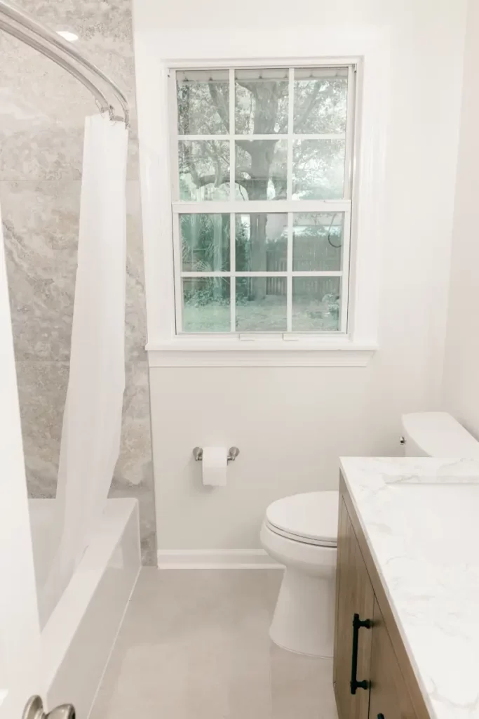 Bathroom interior with white bathtub, grey marble surround tile, gridded window, toilet, and wood vanity in Jacksonville, FL.