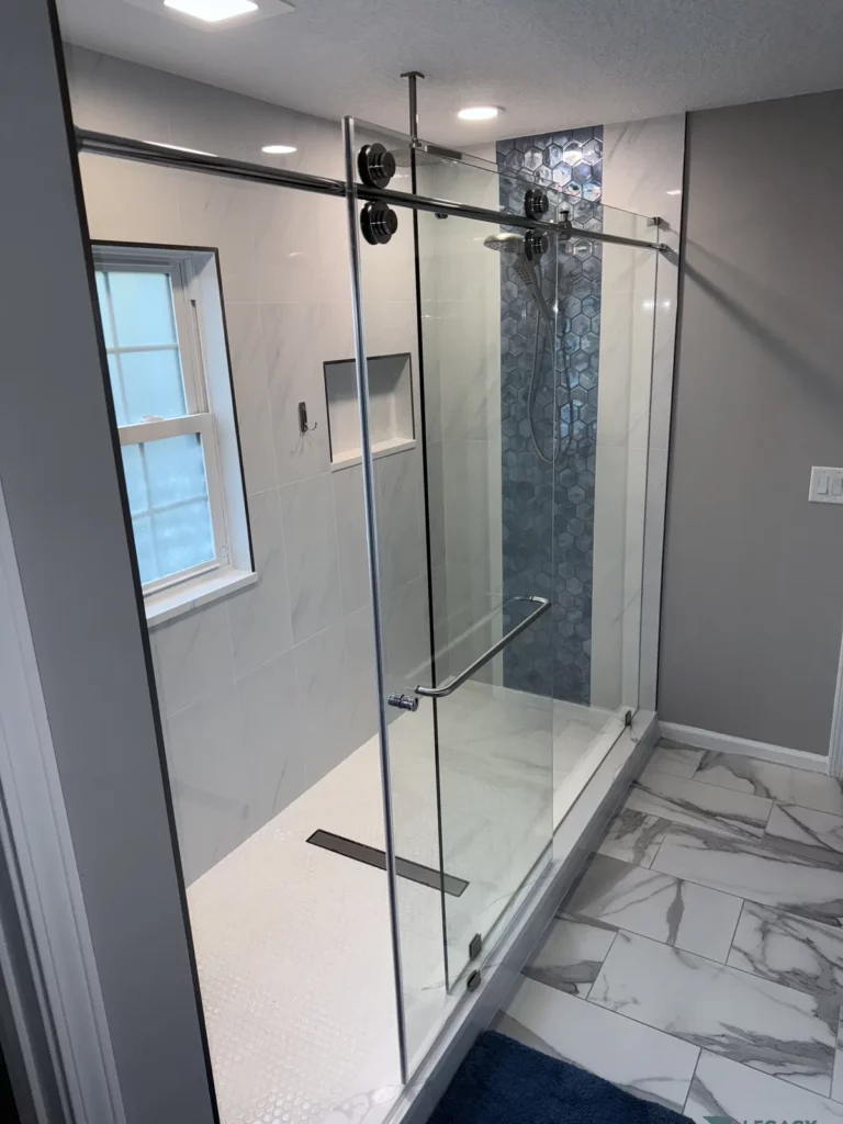 Glass-enclosed walk-in shower with blue hexagon tile, recessed niche, shower fixtures, window, and marble-pattern floor in Jacksonville, FL