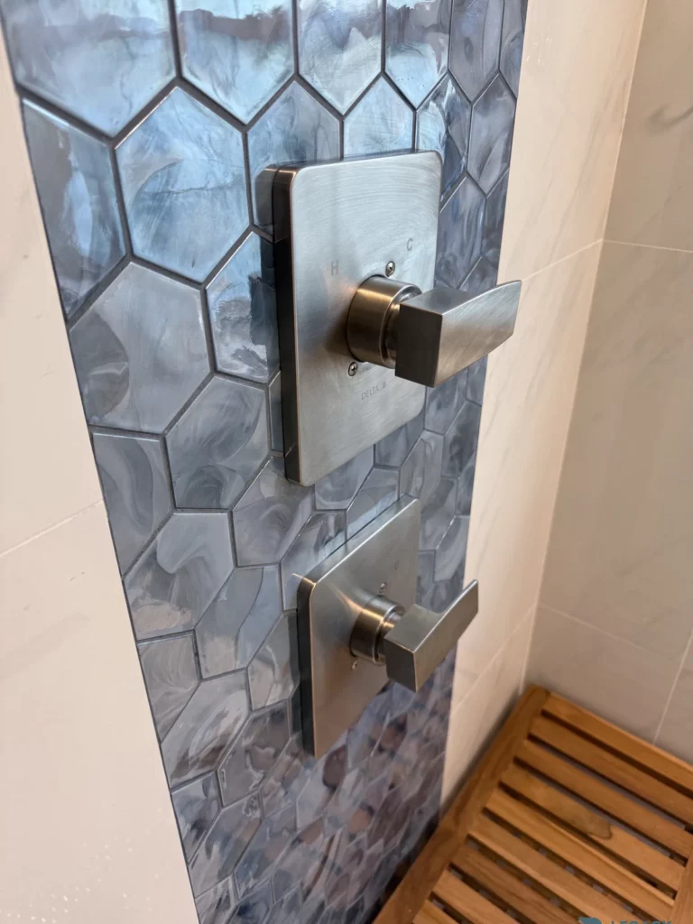 Two brushed metal shower controls mounted on blue marbled hexagon tiles beside a wooden shower bench in Jacksonville, FL