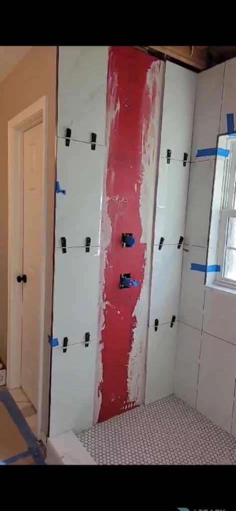 Partially tiled shower with light wall tiles, red waterproofing layer, plumbing controls, window, and hexagon floor tile in Jacksonville, FL