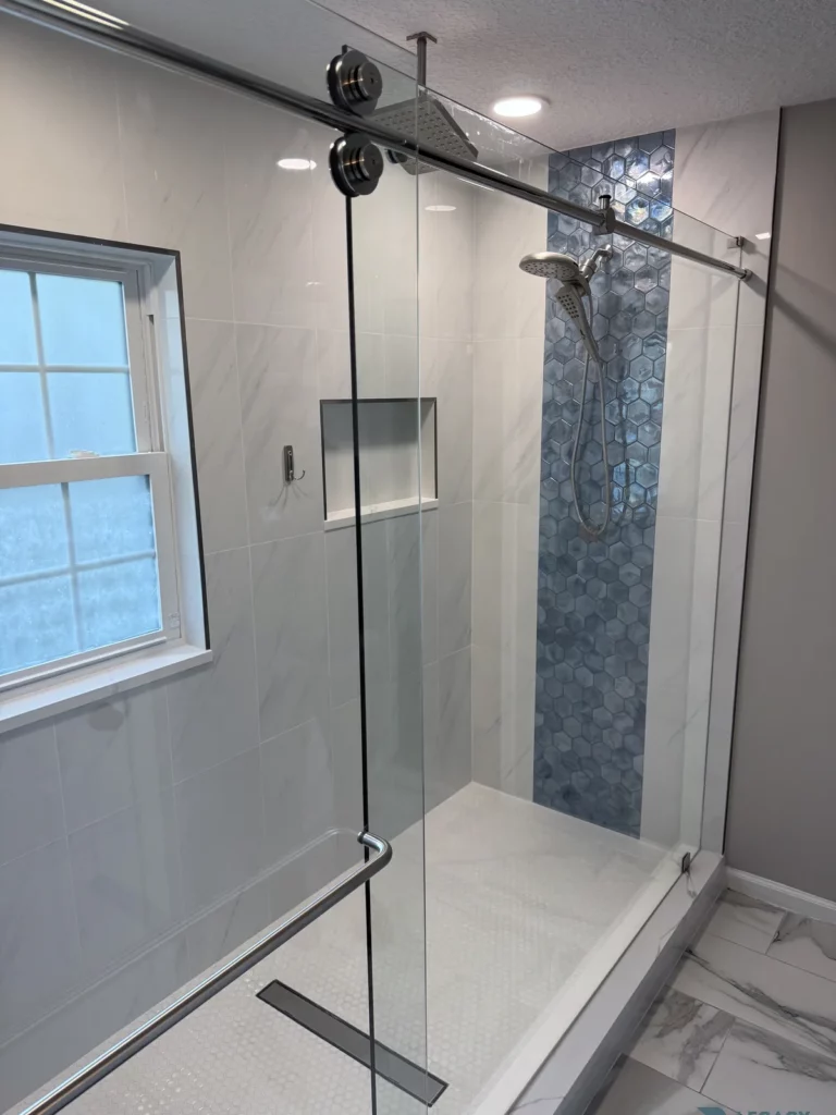 Walk-in shower with sliding glass door, white wall tile, blue hexagon tile accent, recessed niche, window, overhead showerhead, and handheld sprayer.