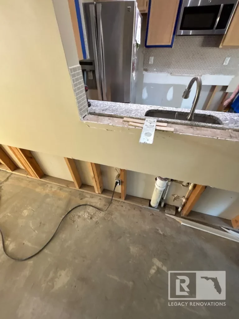 Exposed wooden studs, plumbing pipes, and concrete subfloor under a kitchen pass-through counter during remodeling on Laura Rose Ct.