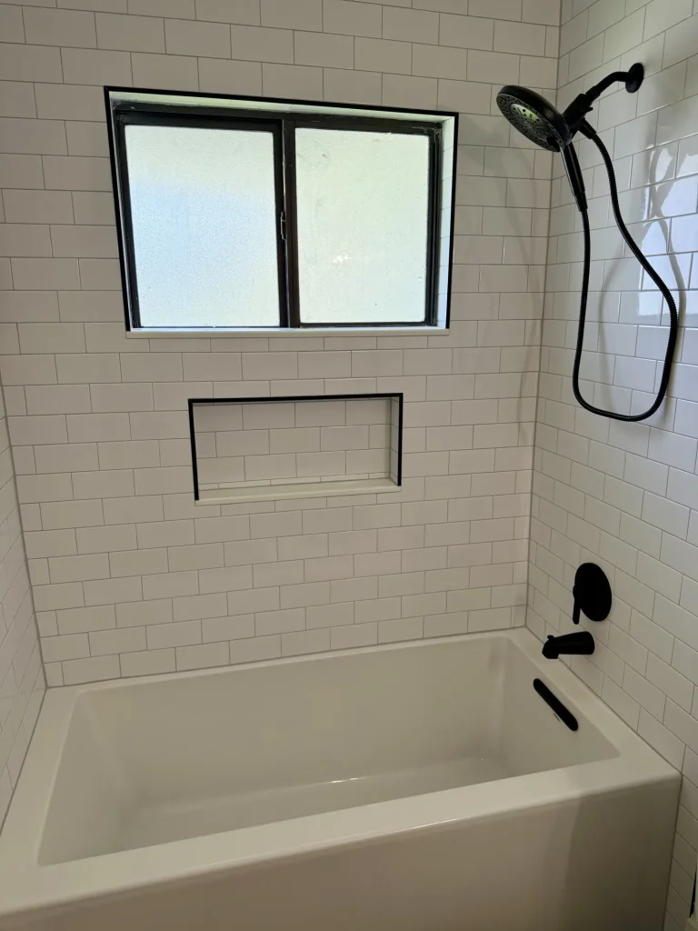 White bathtub beneath a frosted window with white subway tile walls, recessed niche, and black handheld shower fixture in Jacksonville, FL