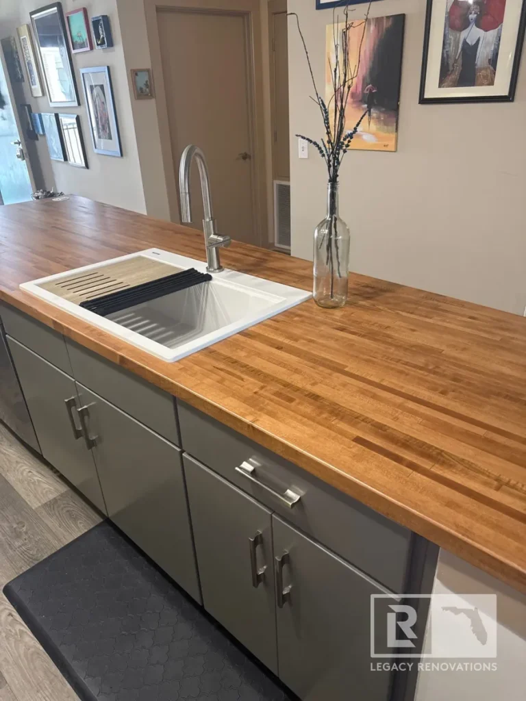 Close-up angle of a wood butcher block countertop featuring an undermount white workstation sink, gooseneck faucet, glass vase decor, grey lower cabinets, and a patterned floor mat.