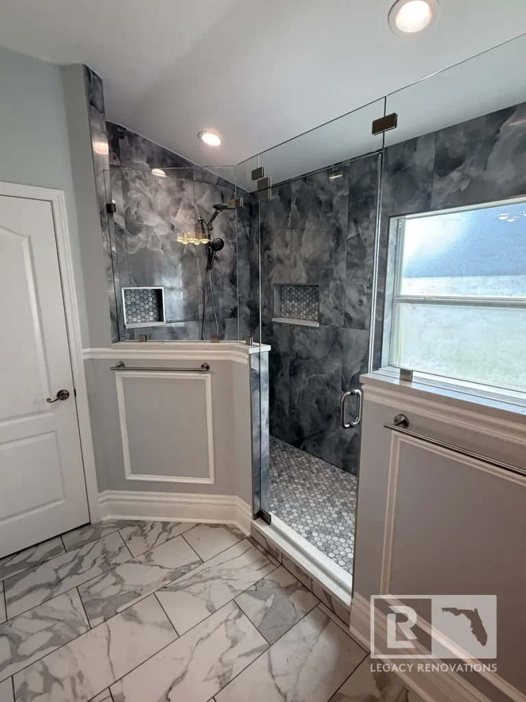 Wide view of a closed frameless glass shower stall framed by grey wainscoting half walls, recessed ceiling lights, white marble-patterned flooring, and a white interior door.