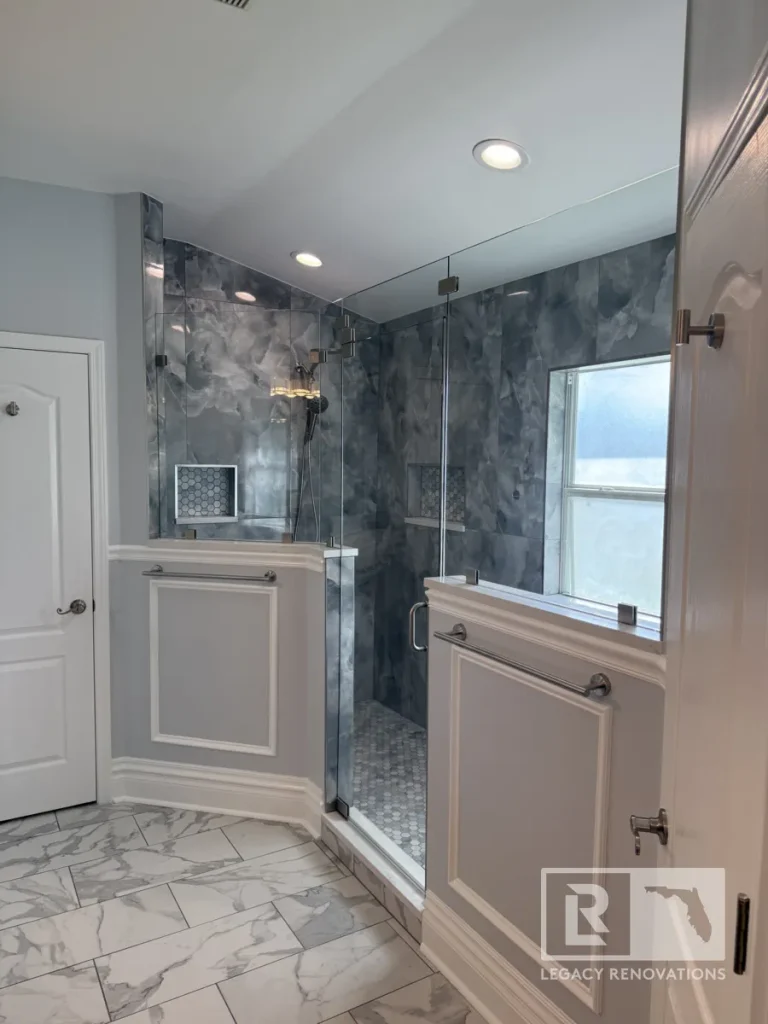 Wide shot showing a corner shower enclosure framed by half walls with wainscoting trim, mounted horizontal towel bars, frameless glass panels, and a open interior doorway.