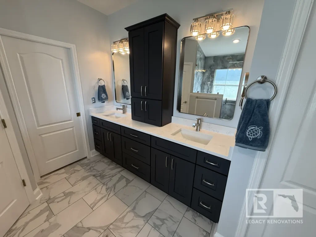 Wide shot of a double-sink navy vanity featuring two undermount basins, twin vanity mirrors with crystal lighting fixtures, a central tall storage cabinet, and white interior doors.