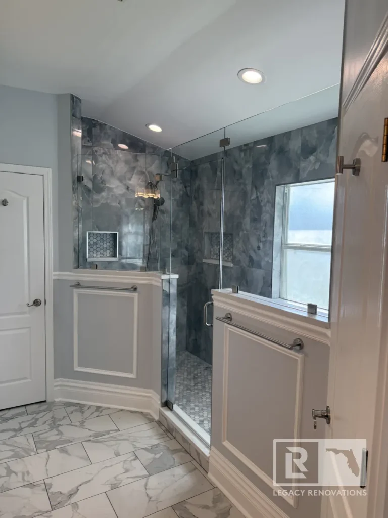 Angle view of a walk-in shower with clear glass upper panels, grey wainscoting lower walls, a closed interior door, recessed ceiling lights, and grey-veined floor tile.