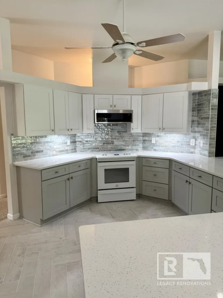 Wide shot of a kitchen featuring a white quartz island foreground, light grey cabinetry, a five-blade wood ceiling fan, glossy brick wall tile, and white appliance range.