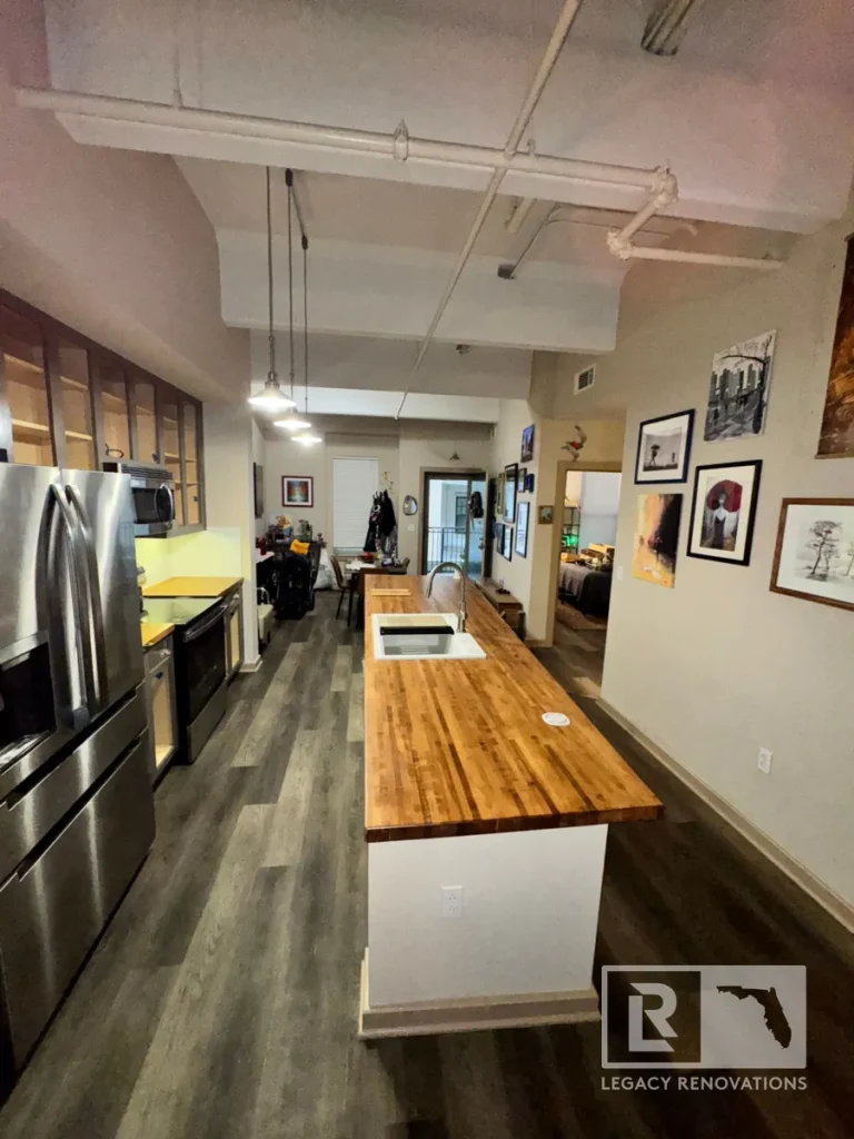 High-angle full shot of an industrial loft kitchen featuring exposed overhead ceiling pipes, a long wood butcher block island with a white sink, stainless steel appliances, and wood-plank flooring.