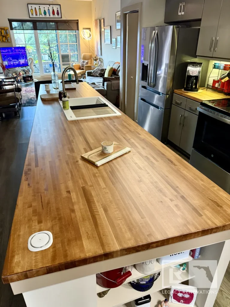 Angle view looking down a finished wood butcher block kitchen island with white undermount sink, marble trivet, and built-in wireless charger, extending toward an open living area with large window and sofa.