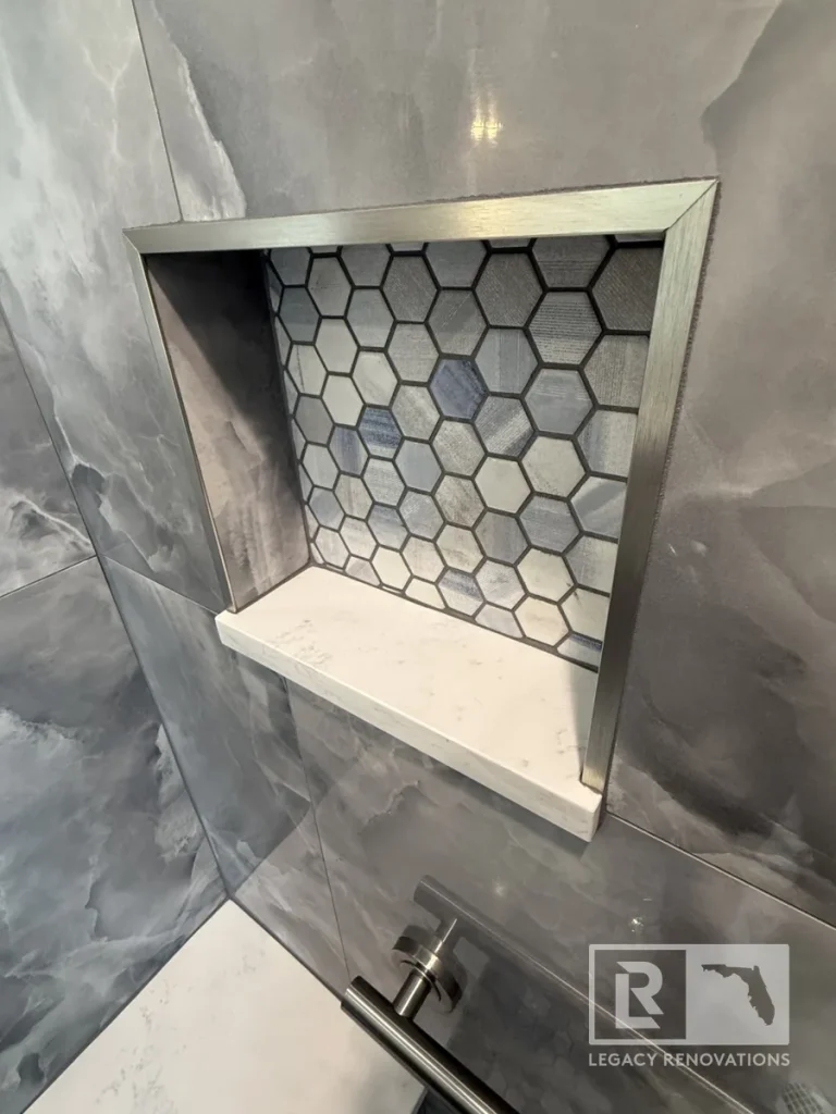 Built-in wall niche framed in brushed nickel trim with blue and grey hexagon mosaic backing tile and a white quartz sill shelf.
