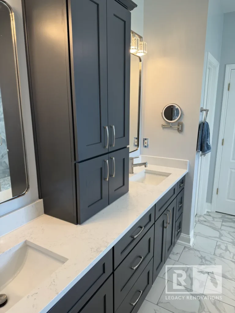 Side-angle view of a navy blue bathroom vanity featuring lower drawers, an upper cabinet tower on a white quartz countertop, two undermount sinks, and a wall-mounted makeup mirror.