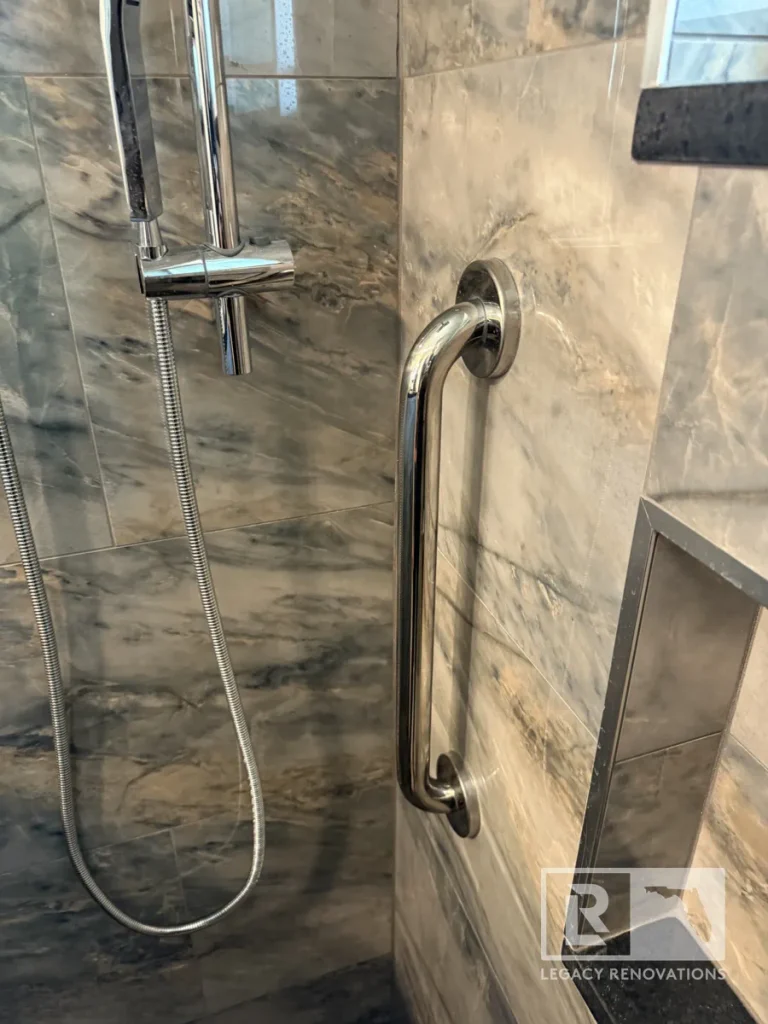 Close-up view of a polished chrome vertical safety grab bar next to a wall slider bar, handheld sprayer hose, niche frame, and grey veined wall tile.