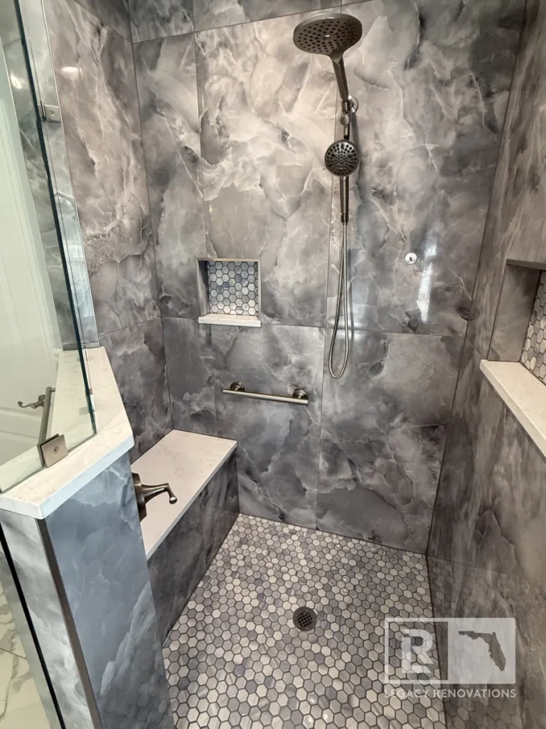 Straight vertical view of a tiled shower stall showing a glass partition wall on a quartz cap, an overhead showerhead assembly, a lower wall water valve, and a hexagonal tile floor.
