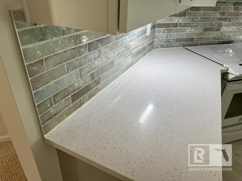 High-angle perspective of a white quartz countertop with grey flecks running beneath hanging wall cabinets and meeting a weathered green-grey brick splash tile.