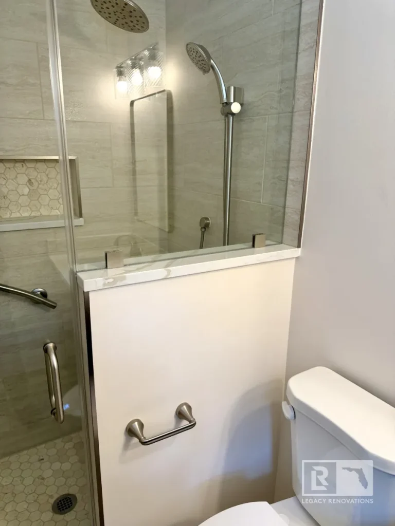 Glass shower panel mounted on a white pony wall with a quartz ledge, showing a dual showerhead setup and handheld wand beside a white toilet.