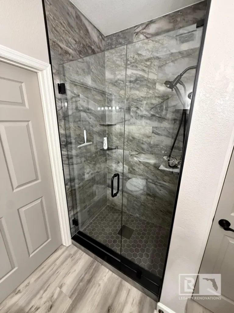 Glass-enclosed shower with gray stone-patterned walls, hexagon floor tiles, black fixtures, and corner shelves in Jacksonville, FL