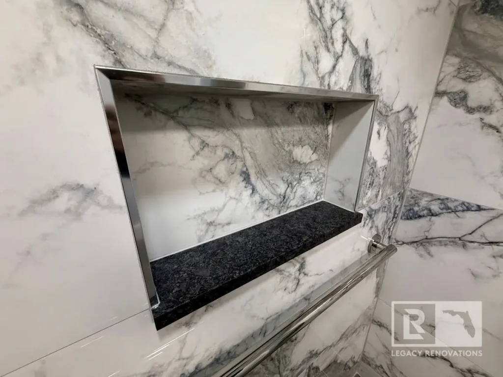 Recessed shower niche with stainless trim, black stone shelf, and marble-look porcelain tile in a custom Jacksonville, FL bathroom remodel.
