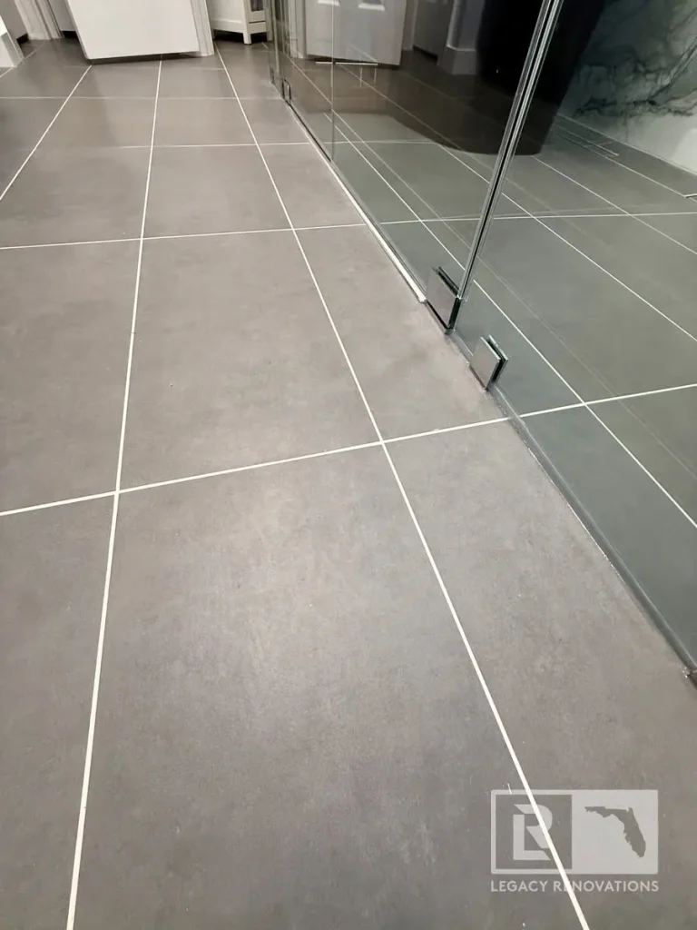 Large-format gray porcelain floor tile with clean grout lines beside a frameless glass shower enclosure in a Jacksonville, FL bathroom remodel.