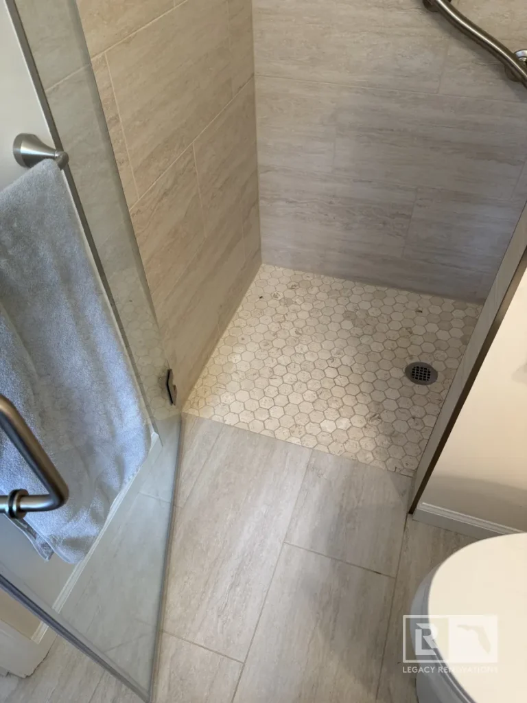 Walk-in shower featuring beige hexagon floor tiles, large format travertine-look wall tiles, a metal grab bar, and a glass door with a grey towel.
