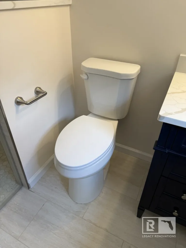 White two-piece toilet positioned on light grey floor tiles between a white interior partition wall and a dark navy blue vanity with a marble countertop.