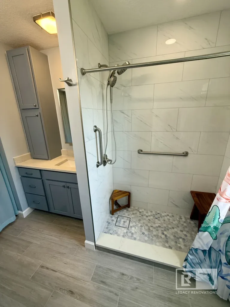 Modern accessible bathroom featuring a walk-in shower with grab bars, pebble mosaic floor, built-in bench, gray vanity, and marble-look wall tile.