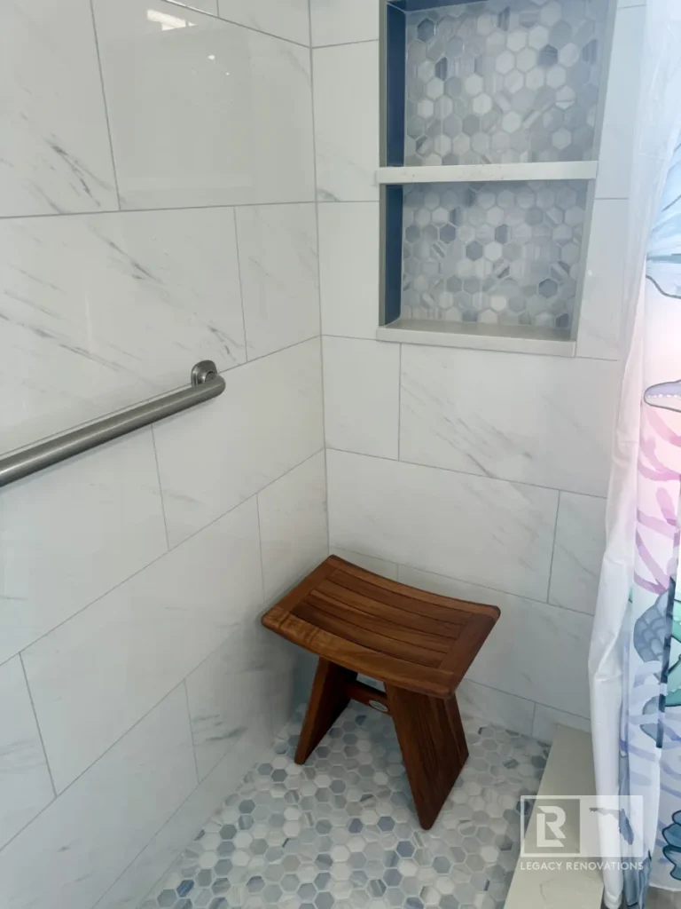 Accessible walk-in shower with marble-look tile, grab bar, teak shower stool, hexagon mosaic floor, and built-in storage niche in a Jacksonville, FL bathroom remodel.