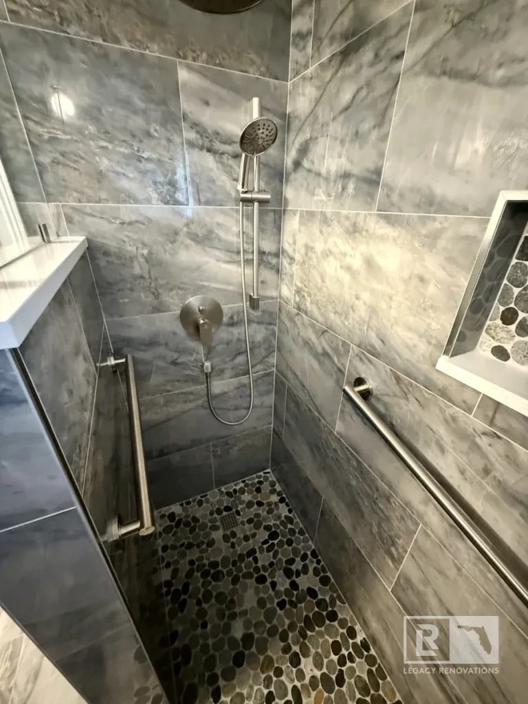 Accessible walk-in shower with gray marble-look tile, pebble stone shower floor, grab bars, handheld shower, and custom recessed niche in Jacksonville, FL.
