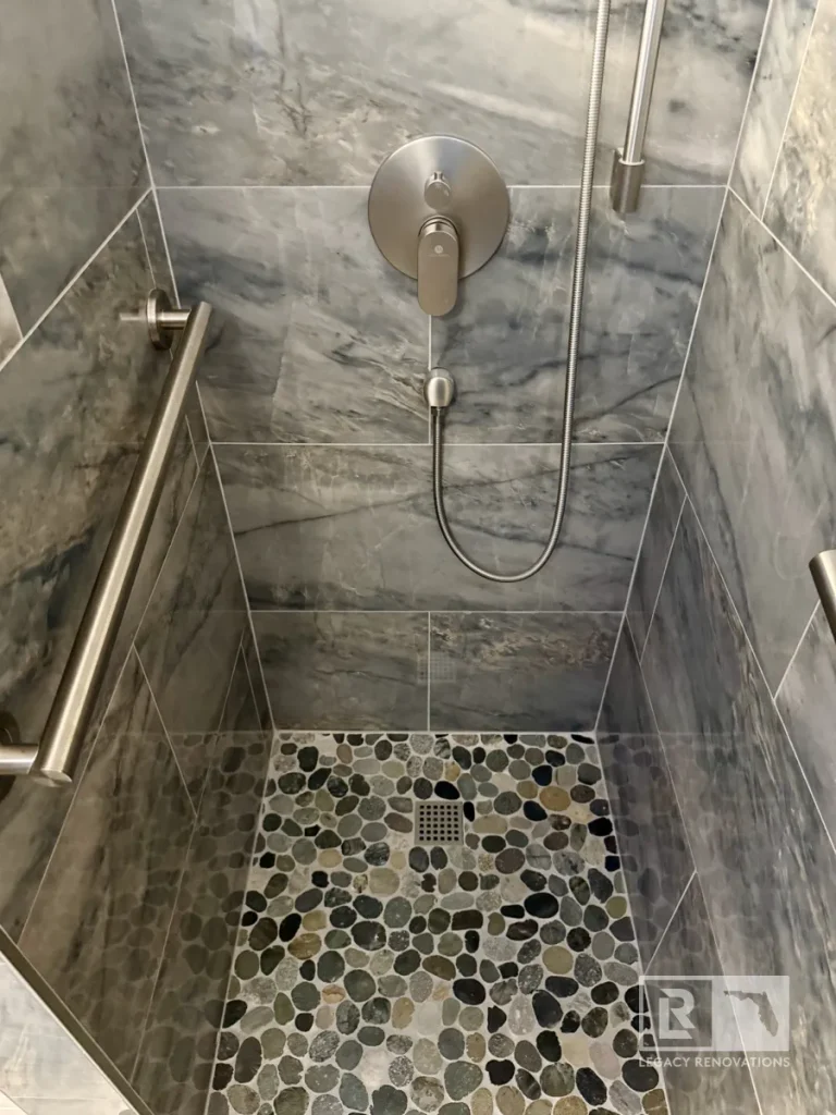 Accessible walk-in shower with gray marble-look tile, pebble stone shower floor, brushed nickel fixtures, grab bars, and a linear drain in Jacksonville, FL.