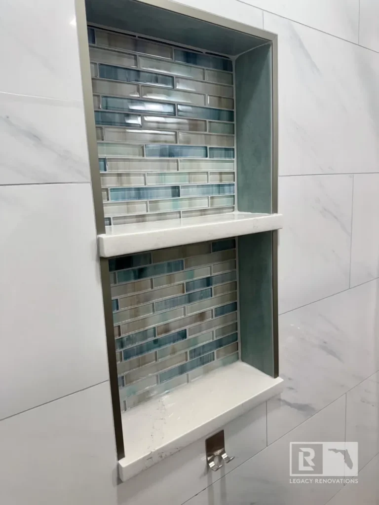 Custom recessed shower niche with blue and gray glass mosaic tile, quartz shelves, and large marble-look wall tile in a Jacksonville, FL bathroom remodel.