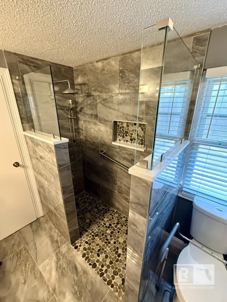 Custom walk-in shower with frameless glass enclosure, gray marble-look tile, pebble shower floor, rainfall showerhead, and recessed niche in Jacksonville, FL.