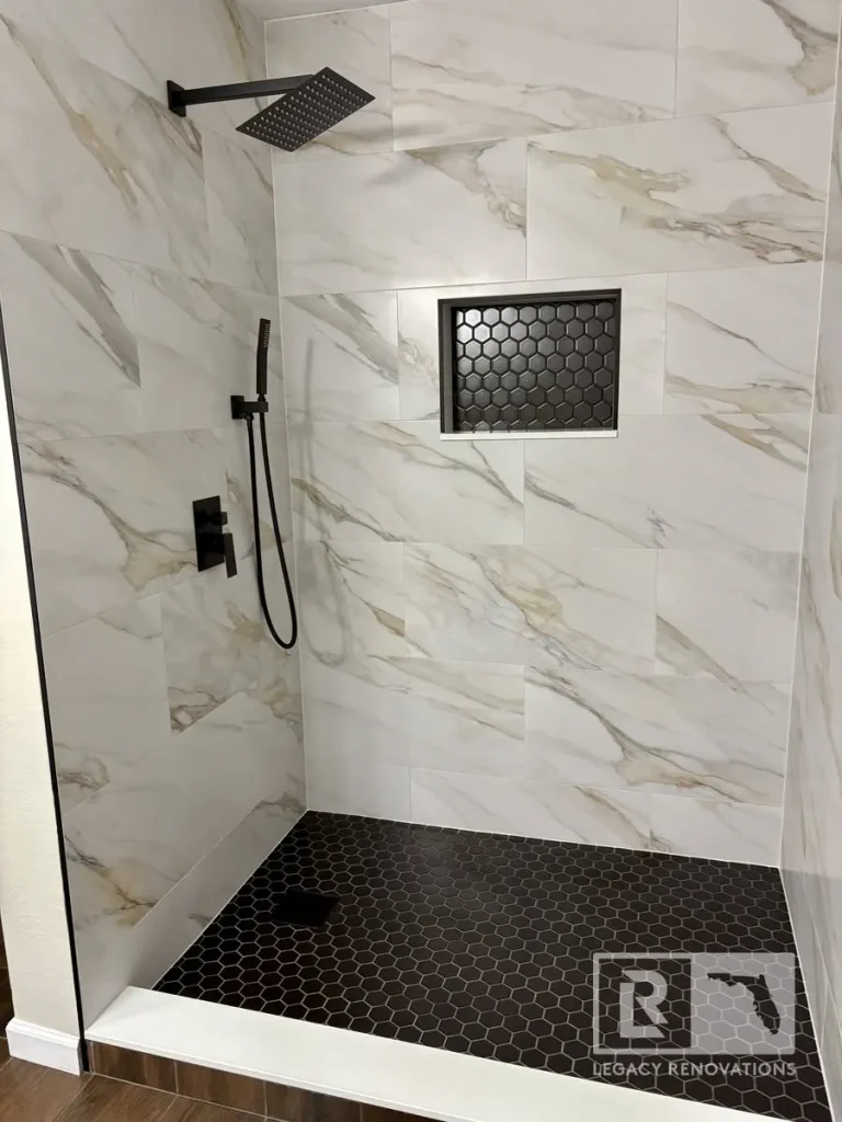 Custom walk-in shower in Jacksonville, FL featuring large-format marble-look tile, black hexagon floor tile, matte black rain shower fixtures, handheld shower, and a recessed shower niche.