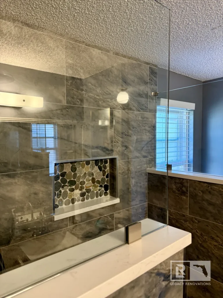 Frameless glass shower with gray marble-look tile, custom pebble stone shower niche, and half wall in a Jacksonville, FL bathroom remodel.