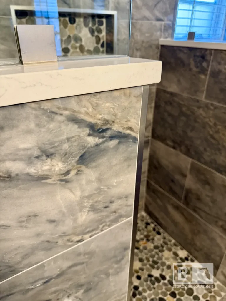 Frameless glass walk-in shower with gray marble-look porcelain tile, quartz curb, and pebble shower floor in a Jacksonville, FL bathroom remodel.