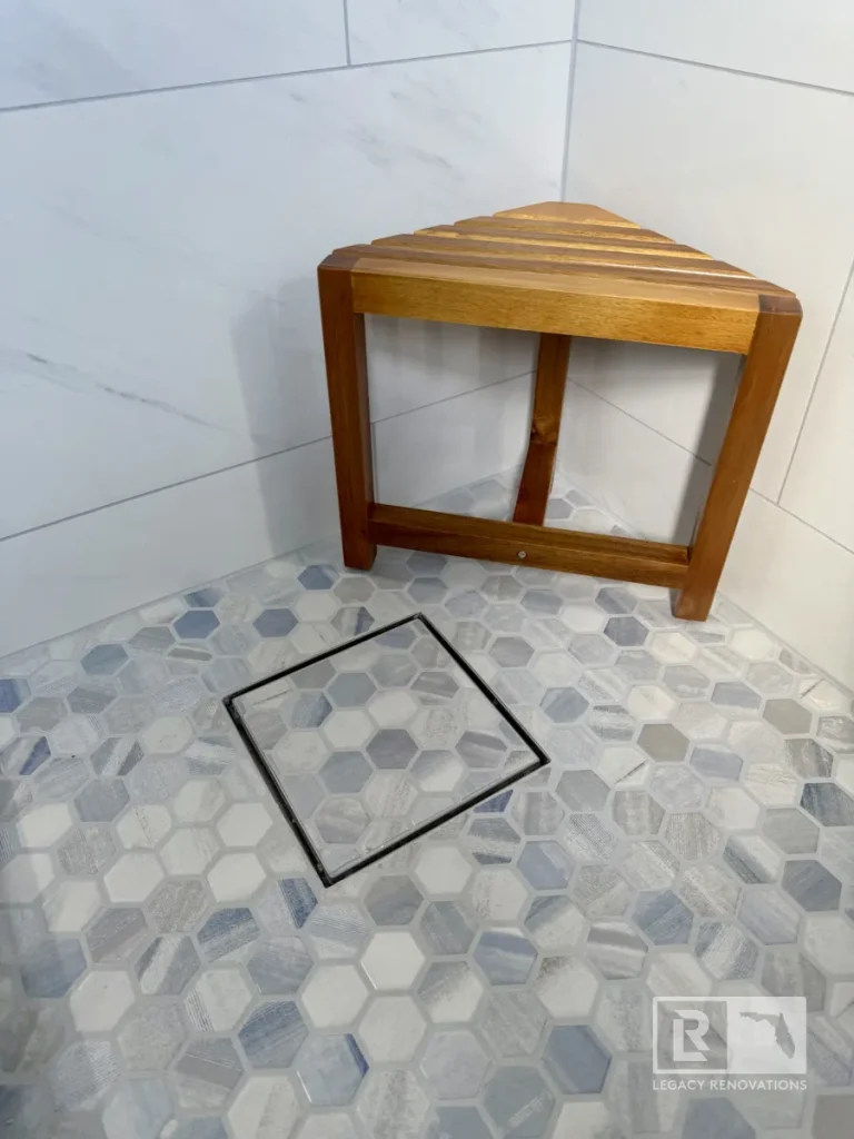 Close-up of a walk-in shower featuring gray and white hexagon mosaic floor tile, a recessed shower drain, and a teak corner stool in a Jacksonville, FL bathroom remodel.