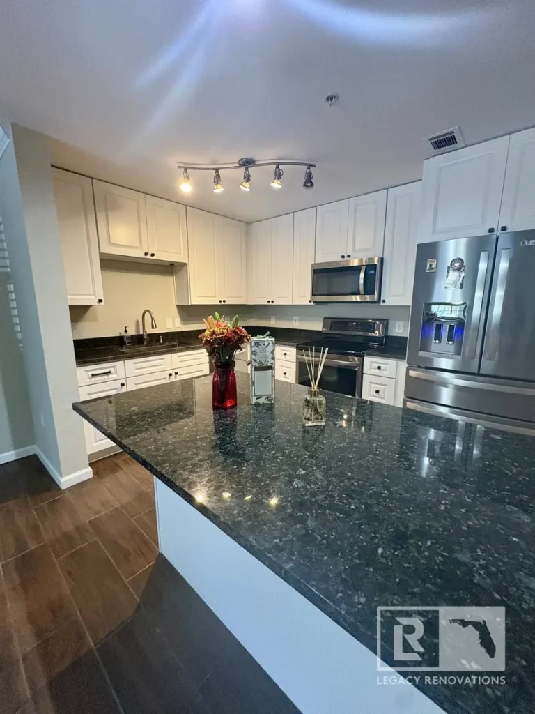 Modern kitchen featuring white shaker cabinets, polished black granite countertops, a large island, stainless steel appliances, and wood-look tile flooring.