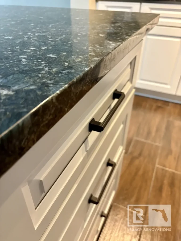 Close-up of white shaker kitchen cabinets with matte black hardware, polished black granite countertops, and wood-look tile flooring.