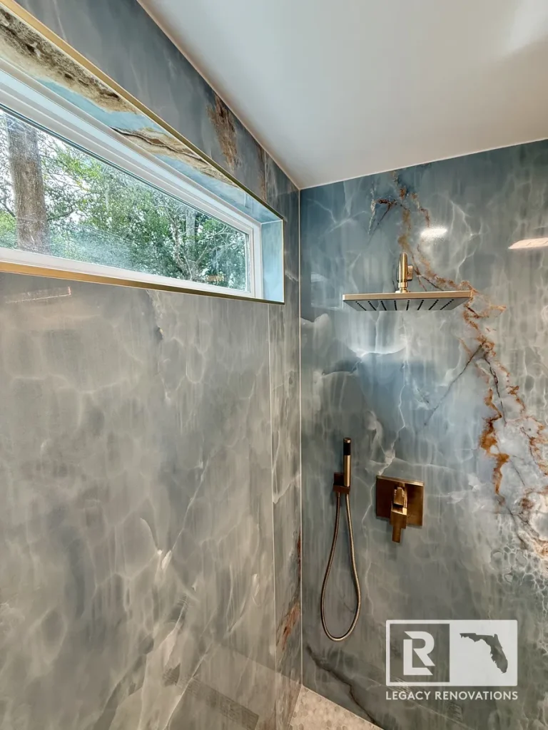 Blue onyx-look walk-in shower with brushed bronze rainfall shower head, handheld sprayer, and transom window in a Jacksonville, FL bathroom remodel