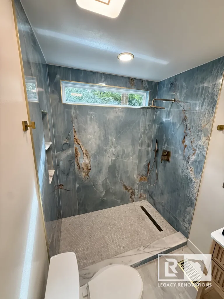 Frameless walk-in shower with large-format porcelain slab walls, rainfall shower system, linear drain, mosaic tile floor, and transom window in a Jacksonville, FL bathroom remodel