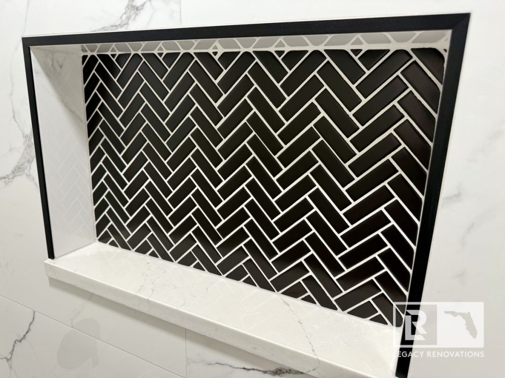 Close-up of a recessed shower niche with black herringbone accent tile, white solid surface shelf, and marble-look porcelain wall tile.